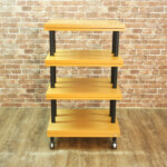 Five-tier wooden rolling cart with black metal supports on a wooden floor against a brick wall, wheels visible at the base.