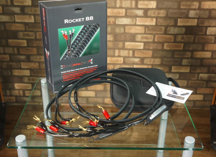 Rocket 88 speaker cable kit on a glass table; black cables with red banana plugs and a retail box in the background against a brick wall.