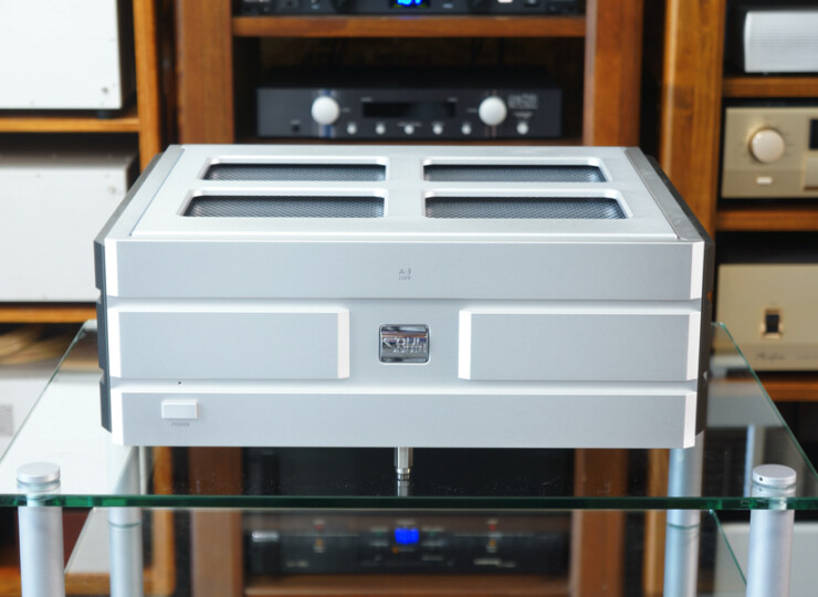 Silver vintage stereo amplifier with vented top on a glass table, surrounded by wooden audio racks behind it.