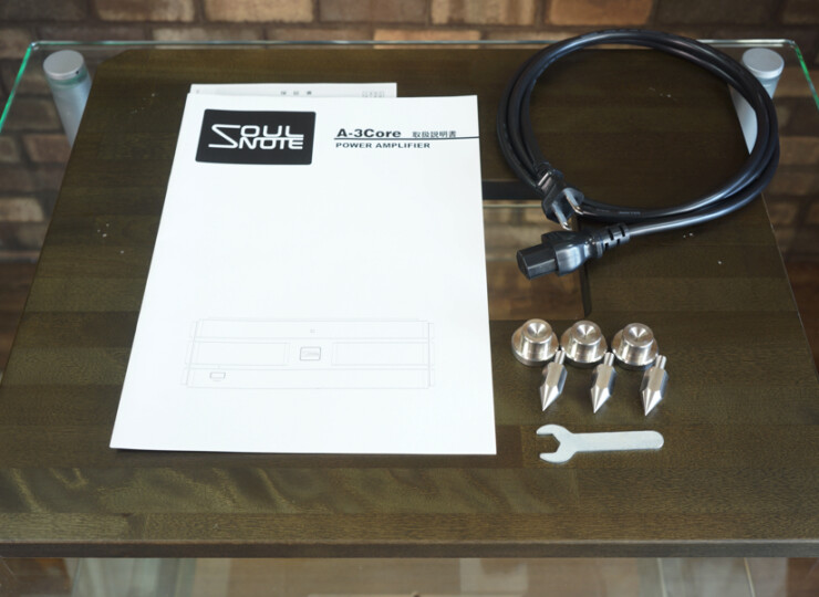 Manual for Soul Note A-3Core power amplifier on a glass-tinished wooden table, with a coiled power cable, four metal fasteners, and a small wrench nearby.