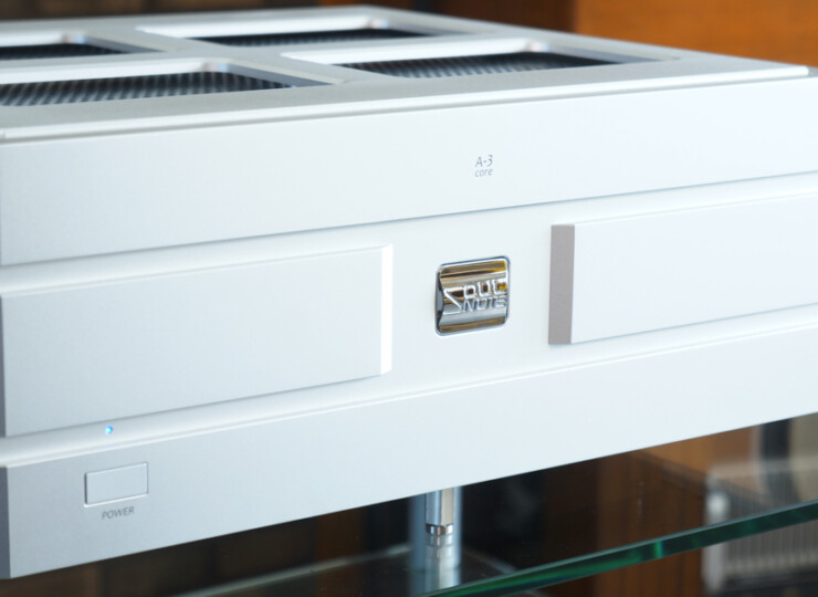 White electronic audio component (likely a vintage amplifier or media chassis) with a chrome badge in the center, mounted on a glass shelf and a small power button on the lower left.