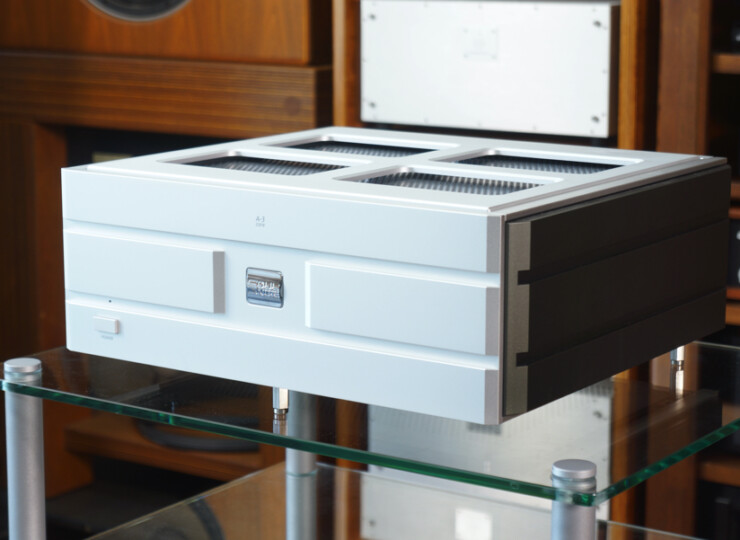 Silver rectangular audio component with a vented top sits on a glass coffee table in a wood-paneled room.