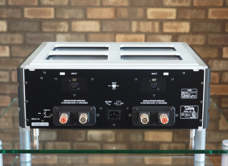 Rear view of a black audio amp showing input connectors, power inlet, and binding posts on a glass shelf against a brick wall.