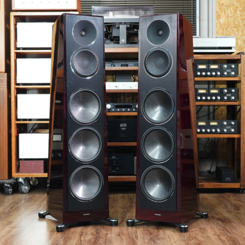 Two large floor-standing speakers with multiple drivers, flanked by wooden audio racks and vintage equipment in a listening room.