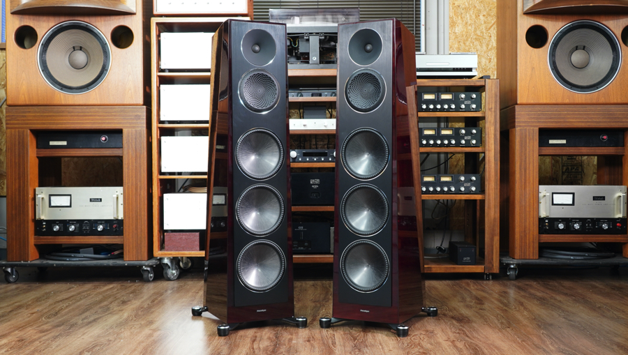 Two large floor-standing speakers with multiple drivers, flanked by wooden audio racks and vintage equipment in a listening room.