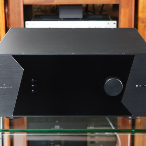 Black stereo amplifier with angular side panels sits on a glass table in front of wooden audio racks and equipment racks in the background.