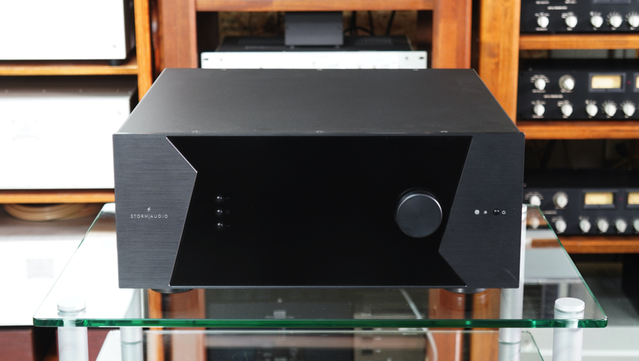 Black stereo amplifier with angular side panels sits on a glass table in front of wooden audio racks and equipment racks in the background.