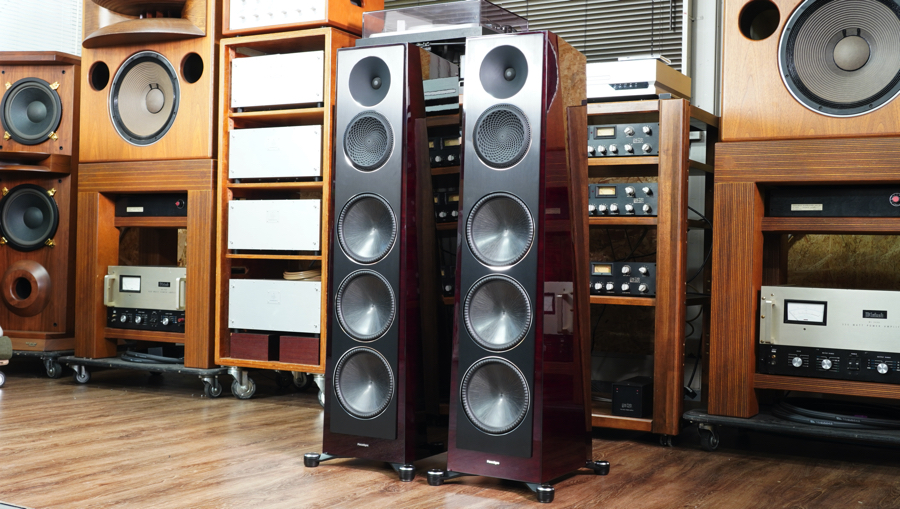 Two tall floor-standing speakers with multiple drivers in a showroom surrounded by vintage audio gear on wooden shelves and floor-to-ceiling equipment racks.