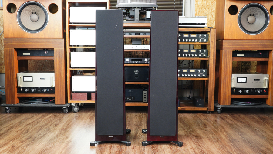 Pair of tall floor-standing speakers in a wood-finished listening room with vintage audio gear behind.