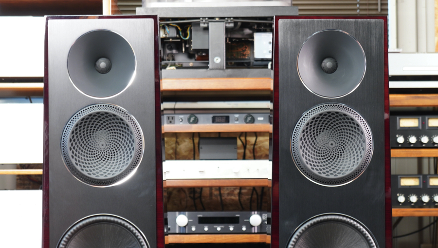 Pair of tall floor-standing speakers with three drivers on each cabinet, flanking an open audio equipment rack with amps and components.