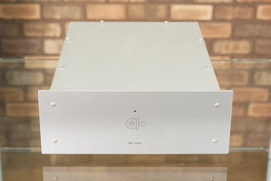 1 | REWIRE WEB SHOP | リワイアー株式会社 Silver, boxy hi-fi component with a minimalist 'ello' logo and 'RMC PHONO' label on the front, placed on a glass surface against a brick wall backdrop.