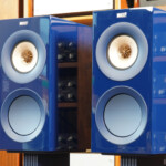 Two blue KEF bookshelf speakers on stands in a room filled with wooden shelves and audio gear.
