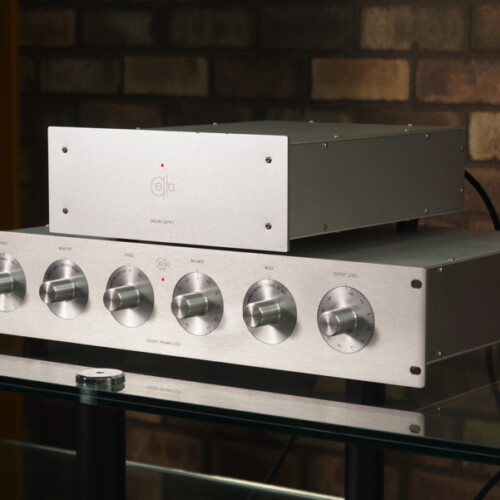 Hi-fi stereo amplifier stack with five large knobs on the brushed aluminum front, placed on a glass shelf against a brick wall.