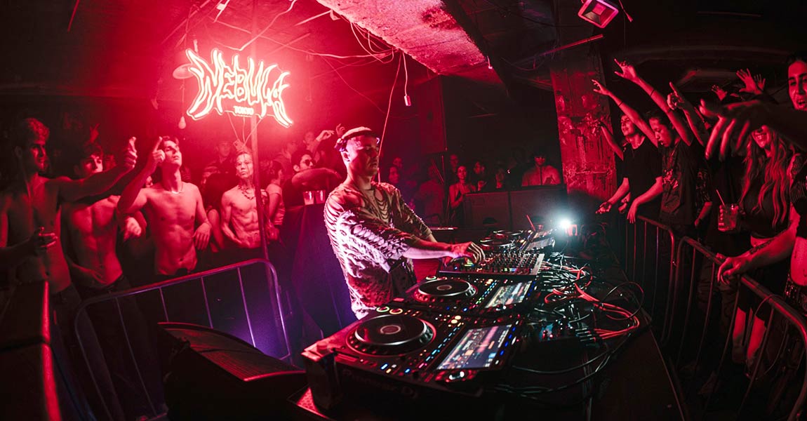 DJ performing at a club surrounded by a cheering crowd, red neon lights, and a mixing console in the foreground.